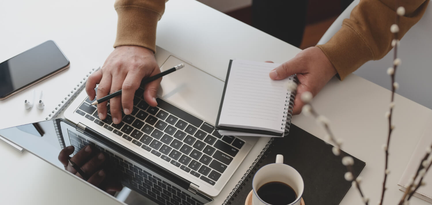 Person using a laptop with one hand typing while holding a pencil and a small notepad in the other hand on a white desk with coffee, phone, and notebooks.