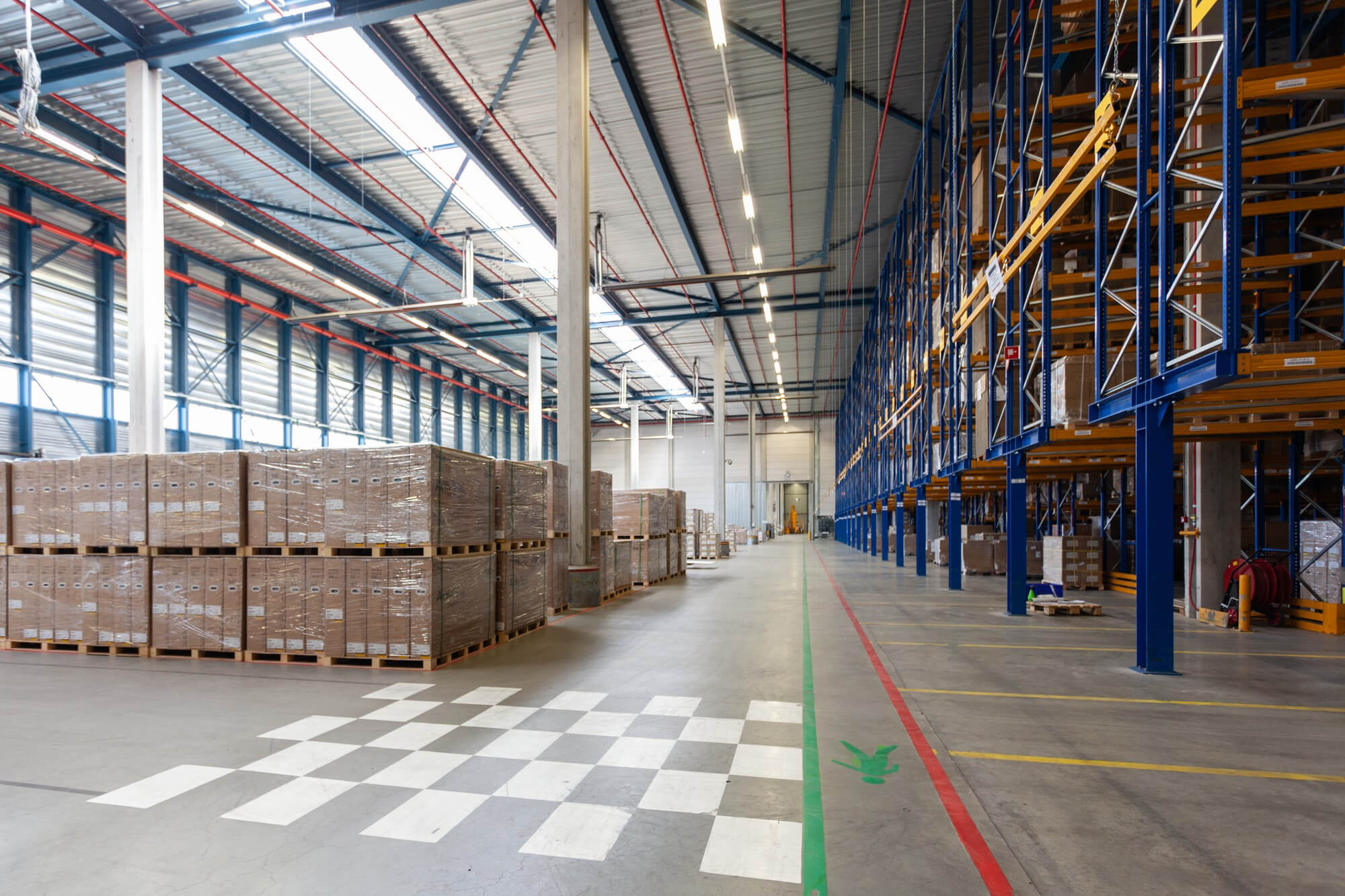 Spacious warehouse interior with stacked pallets on the left and tall blue storage racks on the right under a high ceiling with skylights.