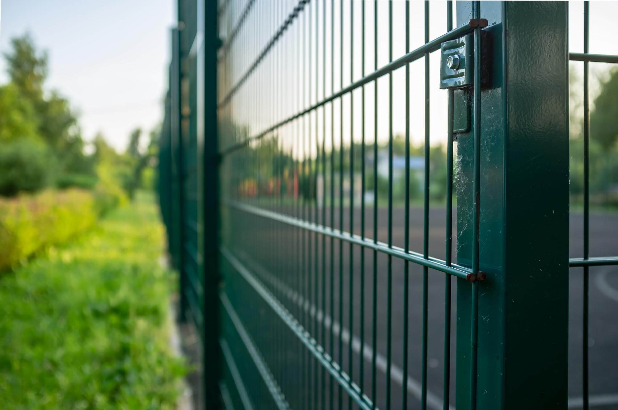 Close-up of a green metal wire fence with blurred grass and trees on the left side and a paved area on the right side.
