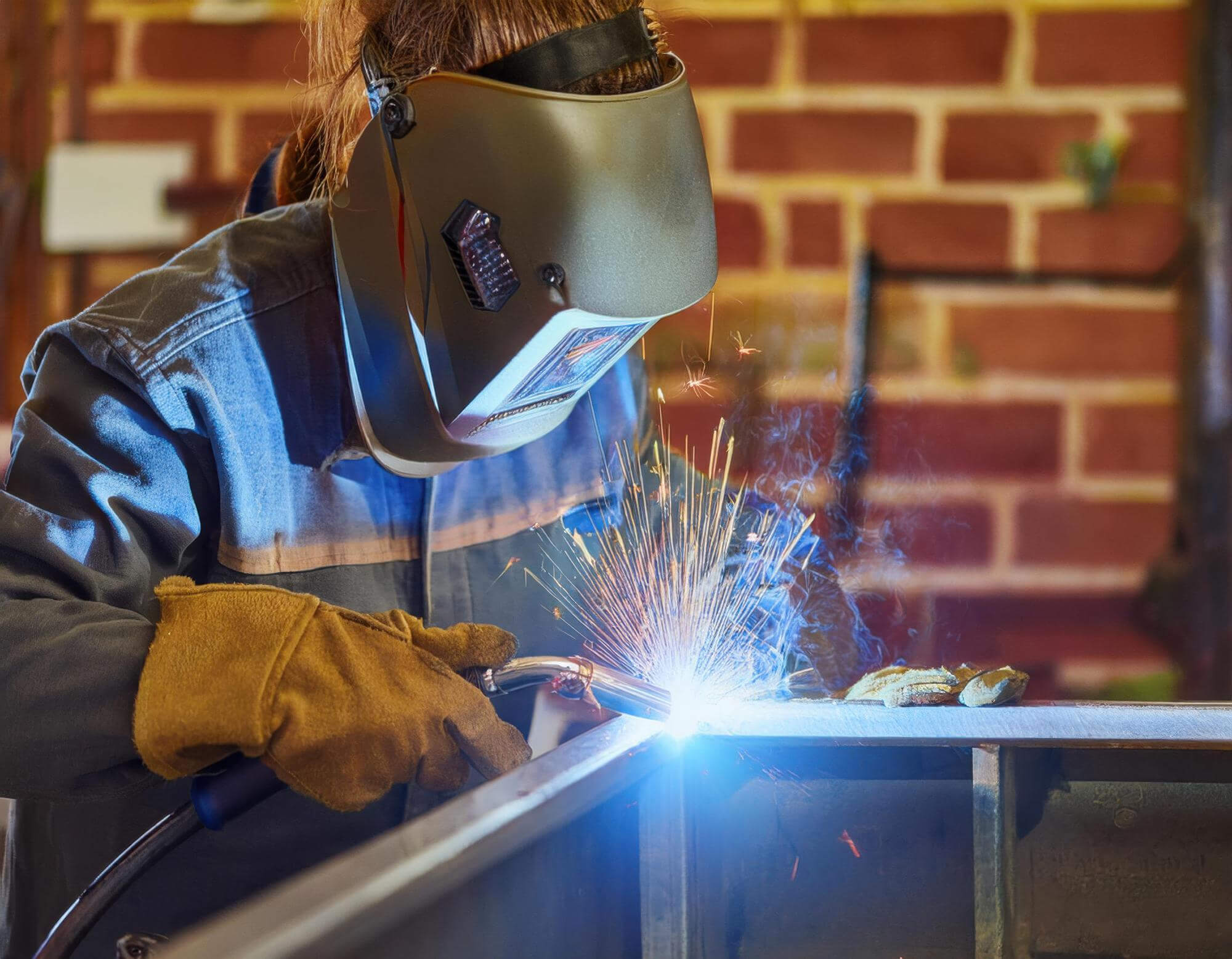 Person wearing protective welding helmet and gloves welding metal with sparks flying.