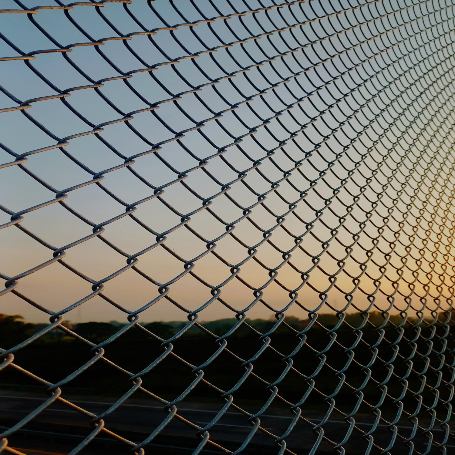 Close-up view of a metal chain-link fence with a sunset sky and landscape in the background.