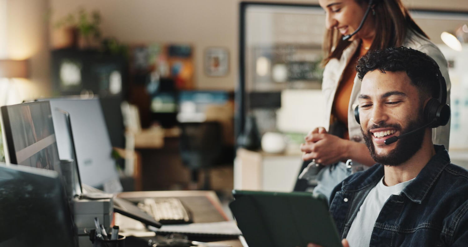Two customer service representatives wearing headsets looking at a tablet in an office.