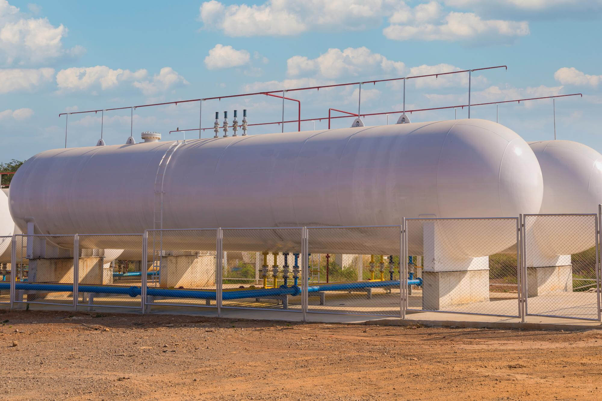 Large industrial horizontal cylindrical storage tanks surrounded by a chain-link fence under a blue sky with clouds.