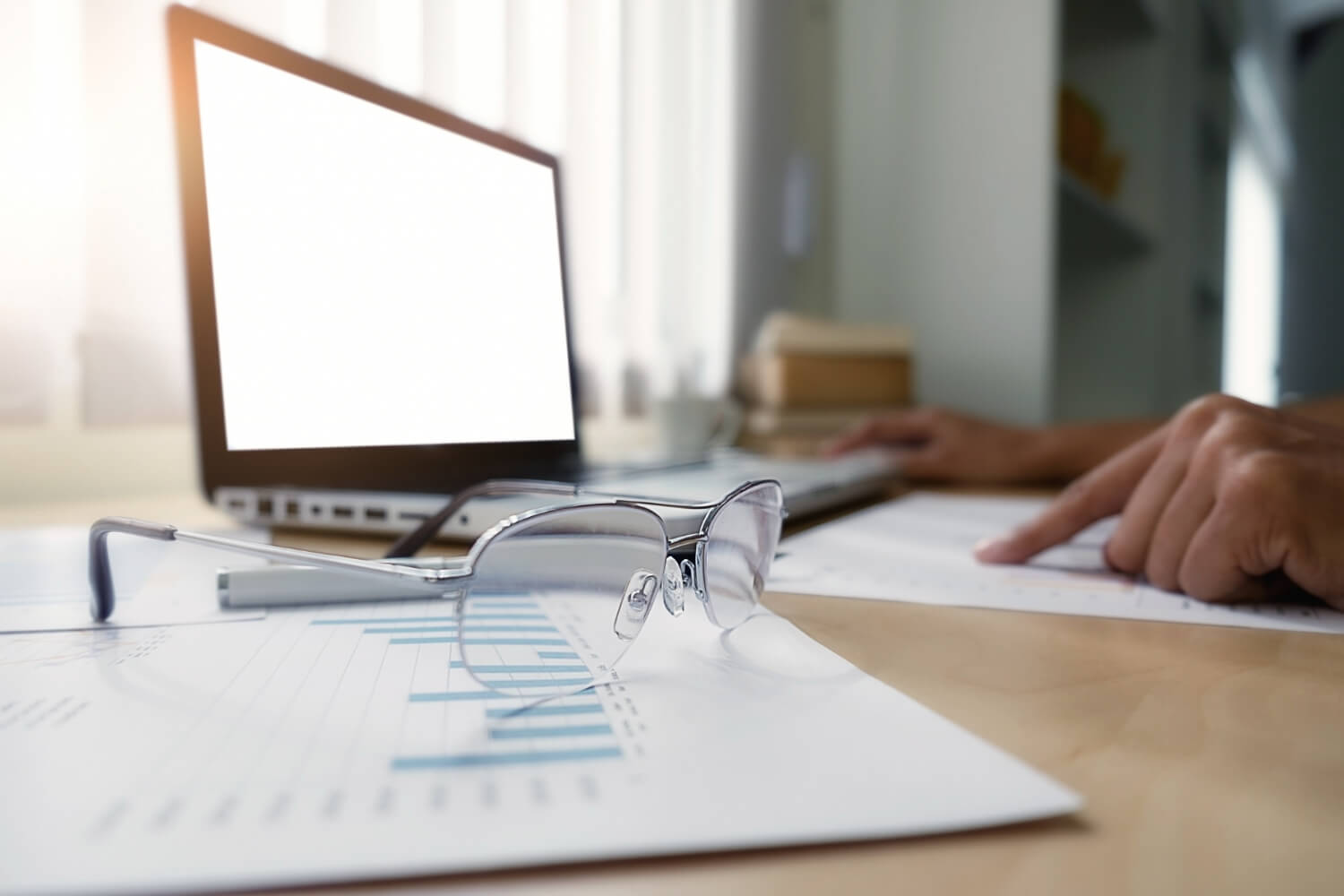 Eyeglasses resting on printed charts with a blurred laptop and person pointing at a paper in the background.