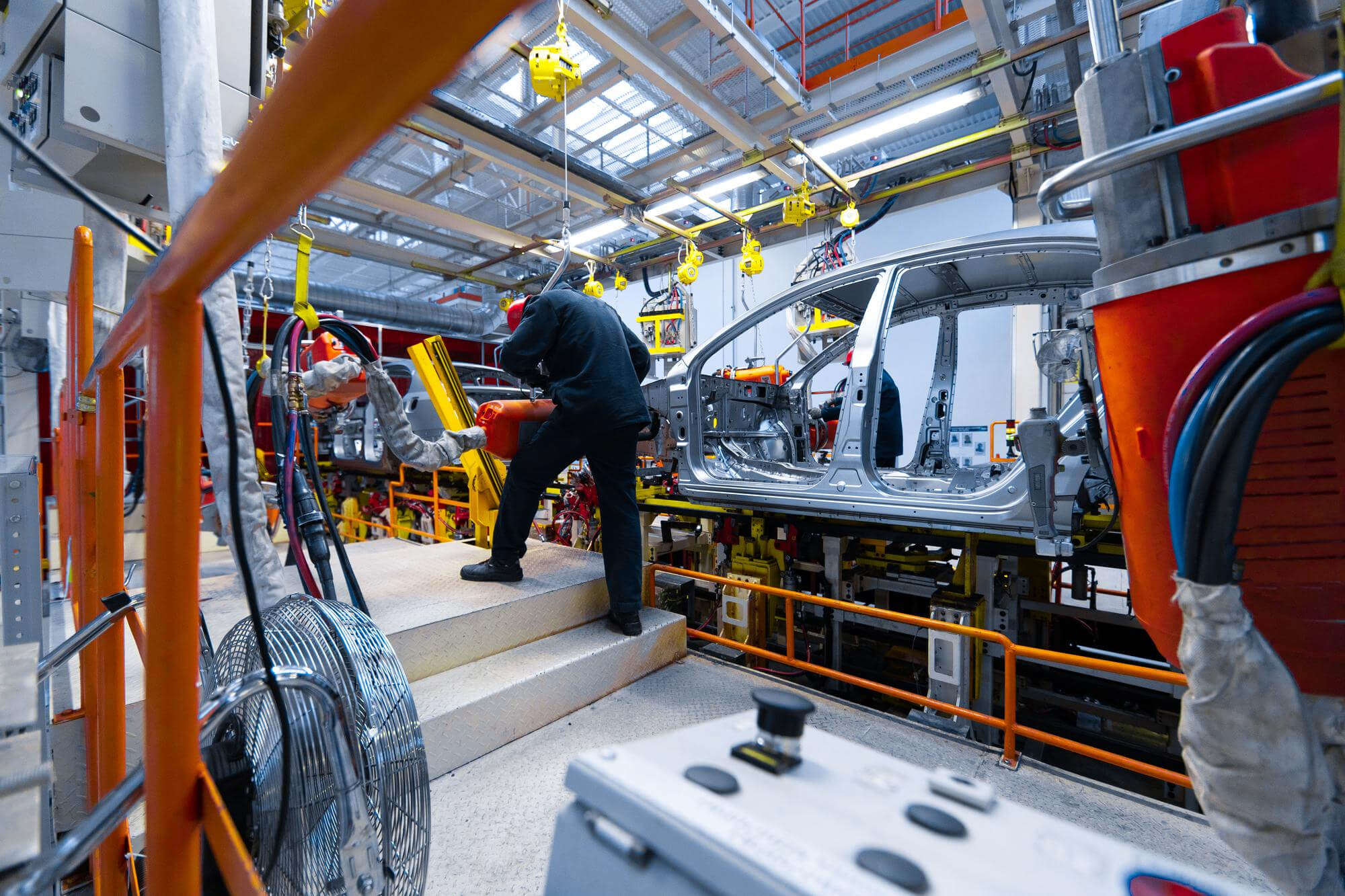 Worker in a red helmet inspecting an automated robotic arm assembling a car frame in a modern factory.