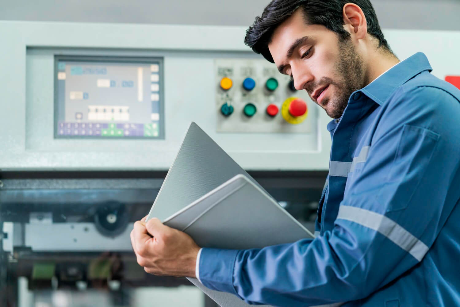 Engineer in blue uniform checking machine settings on an industrial control panel while holding a folder.