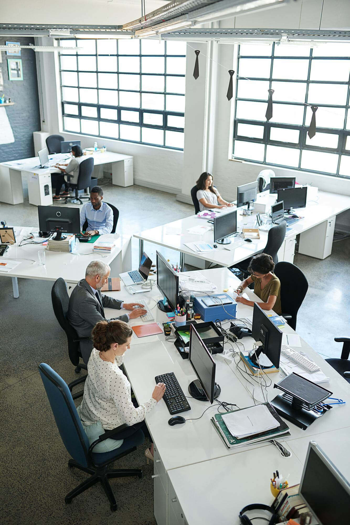 Open office space with diverse colleagues working at desks equipped with computers and office supplies under large windows.
