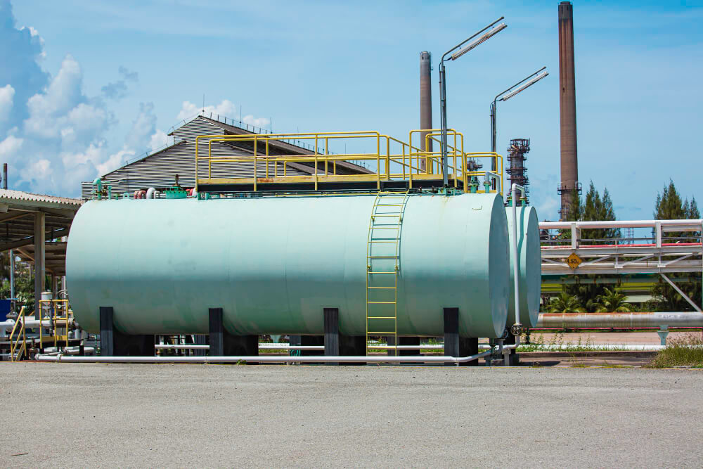 Large light blue horizontal industrial storage tanks with yellow railings and ladder in a factory setting under a blue sky.