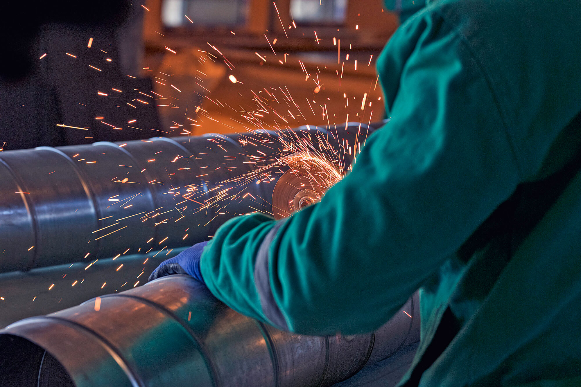 Person in green workwear using an angle grinder to cut a metal pipe, generating bright sparks.