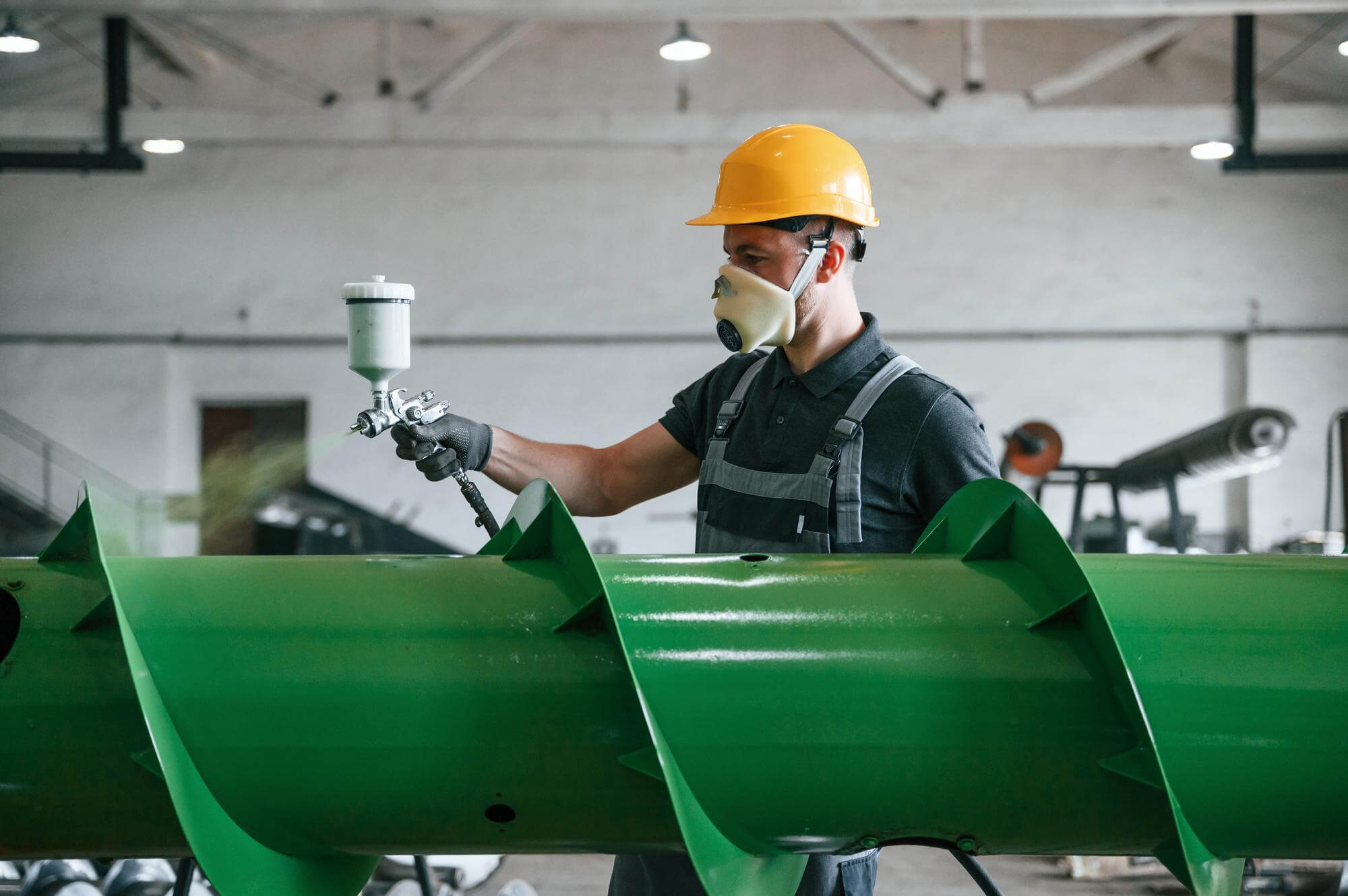 Worker wearing yellow hard hat and face mask spray painting a large green metal industrial auger inside a factory.