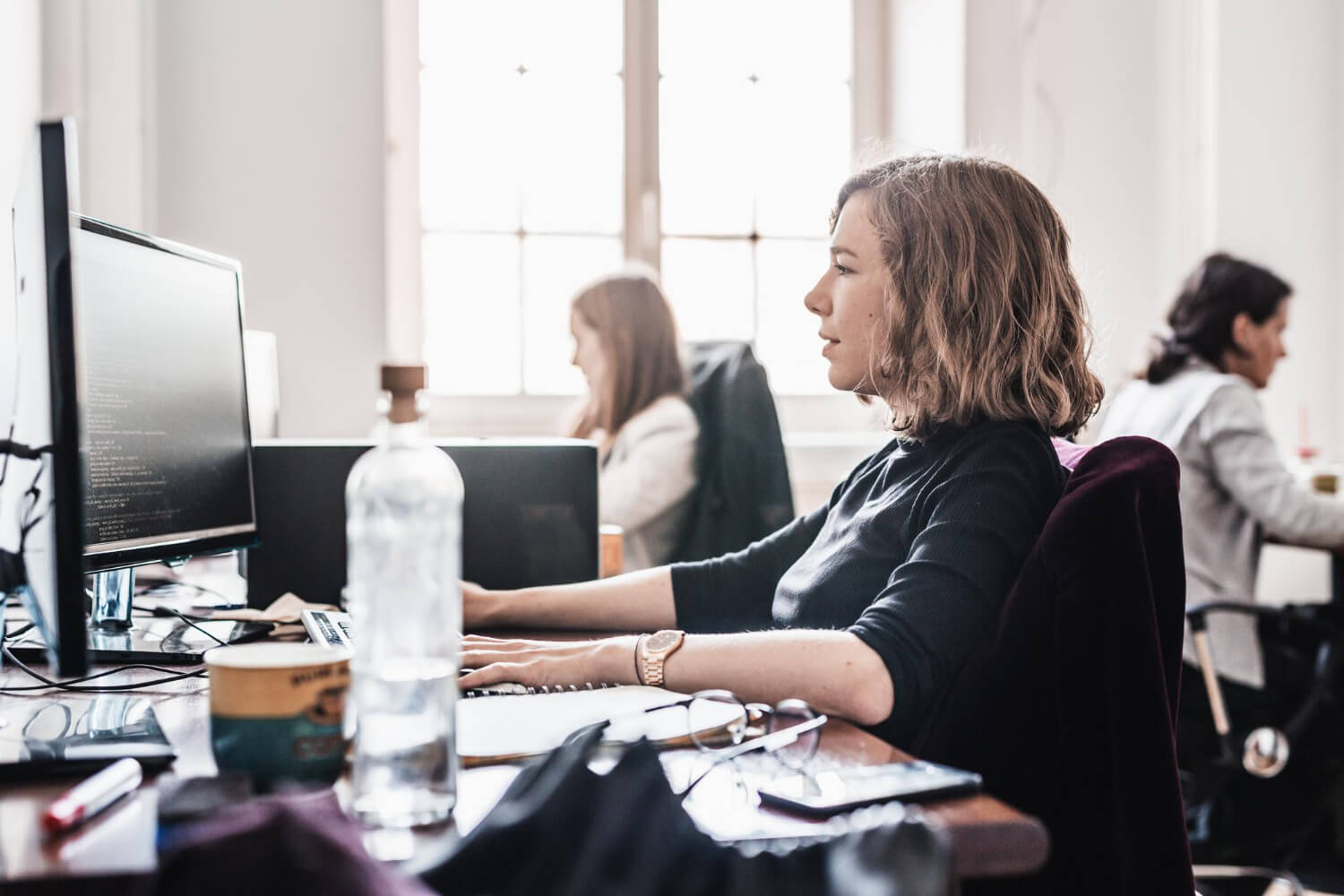 Woman with shoulder-length hair focused on coding at her computer in a busy office with colleagues working in the background.