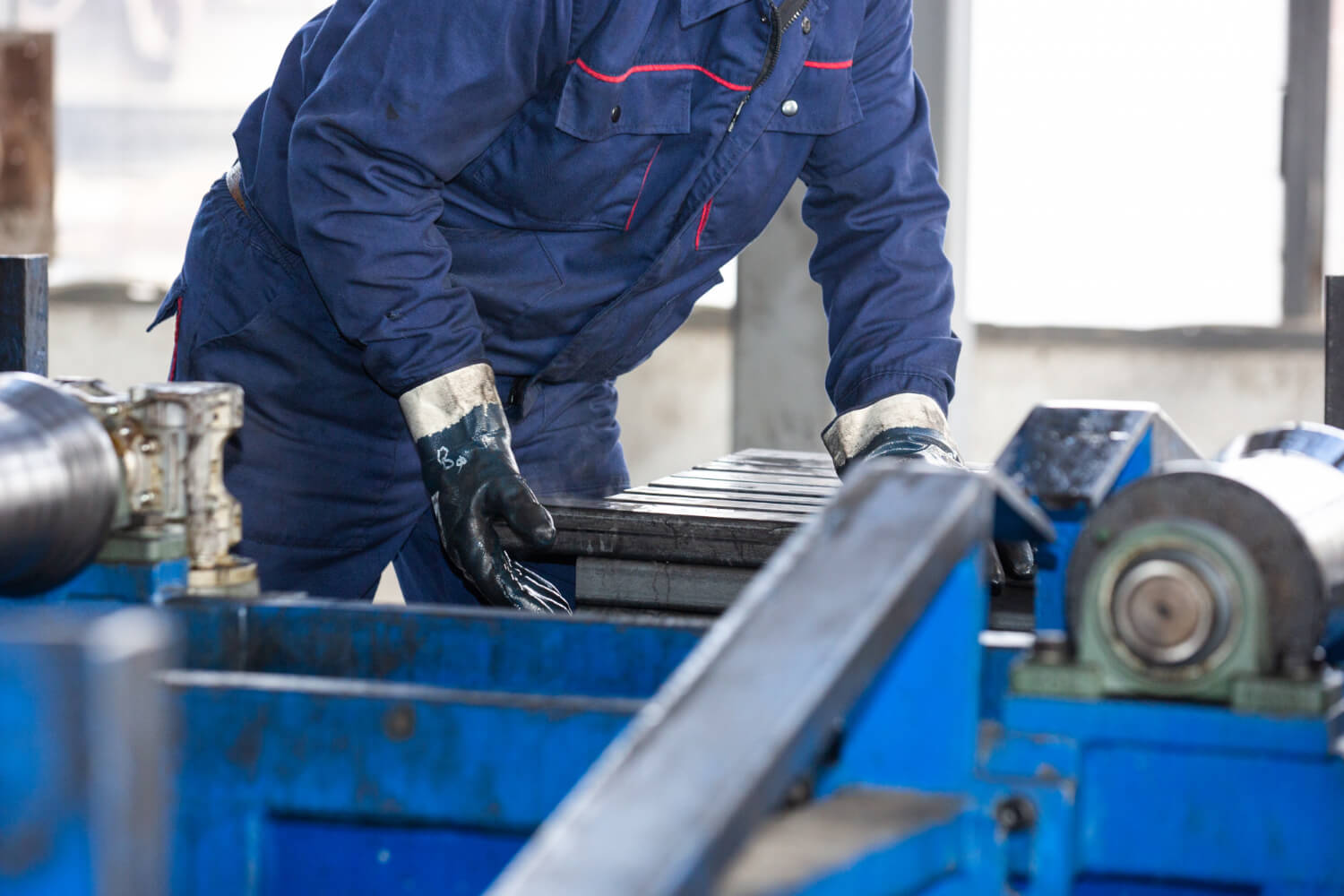 Worker wearing blue coveralls and protective gloves operating blue industrial machinery.