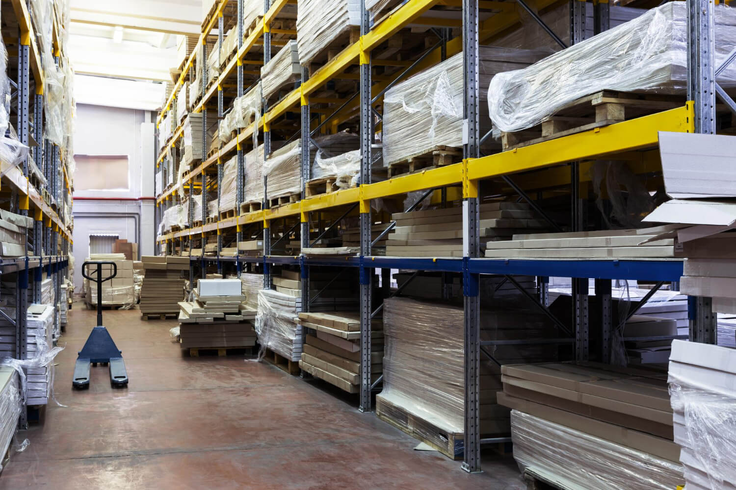 Warehouse aisle with metal shelving stocked with packaged flat panels and a pallet jack on the floor.