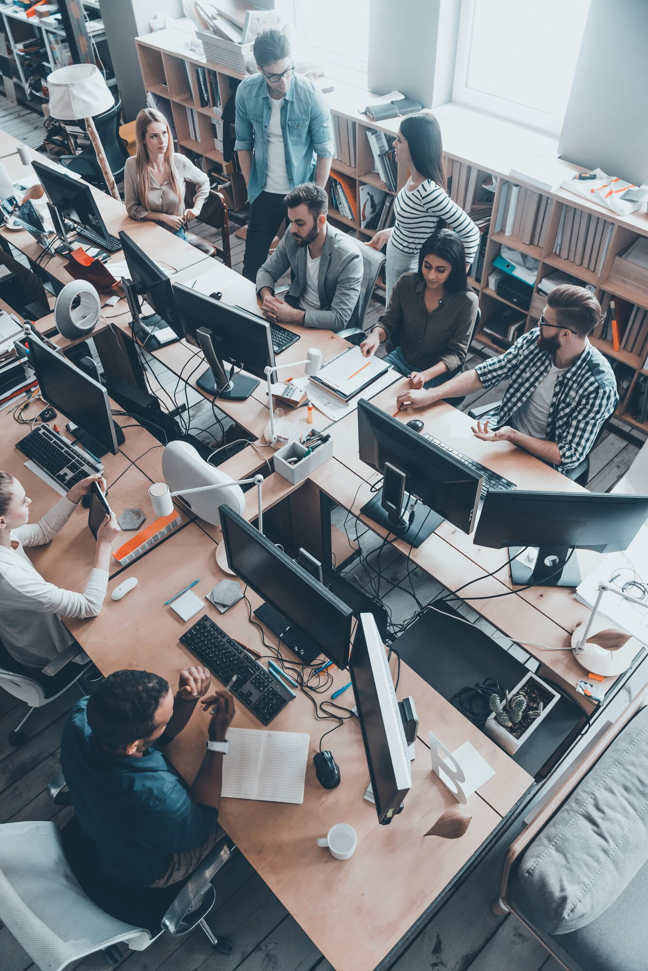 A diverse group of eight coworkers collaborating and discussing around a workspace with multiple computer monitors in a modern office.