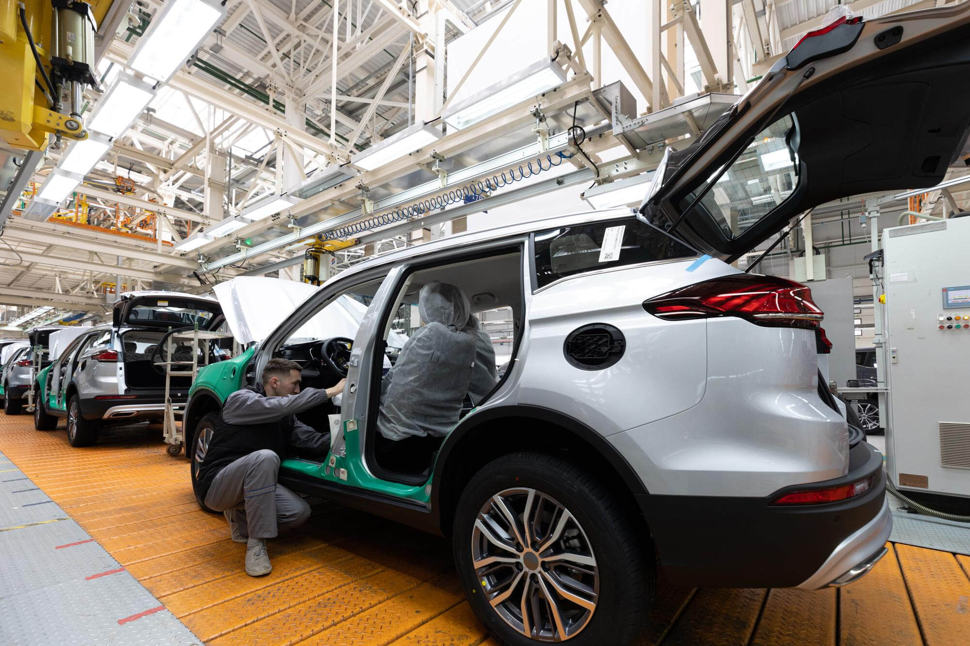 Workers assembling white SUVs on a factory production line with open doors and exposed interiors.