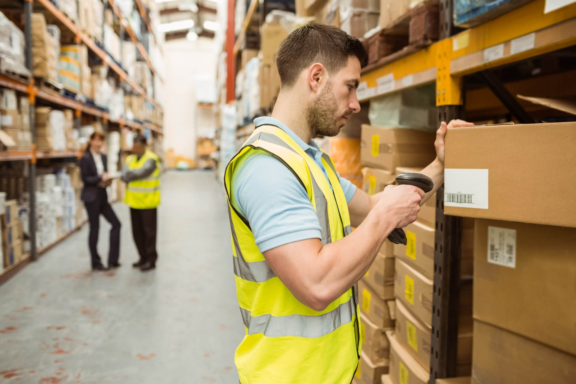 Warehouse worker in a yellow safety vest scanning a barcode on a cardboard box with shelves of packages in the background.
