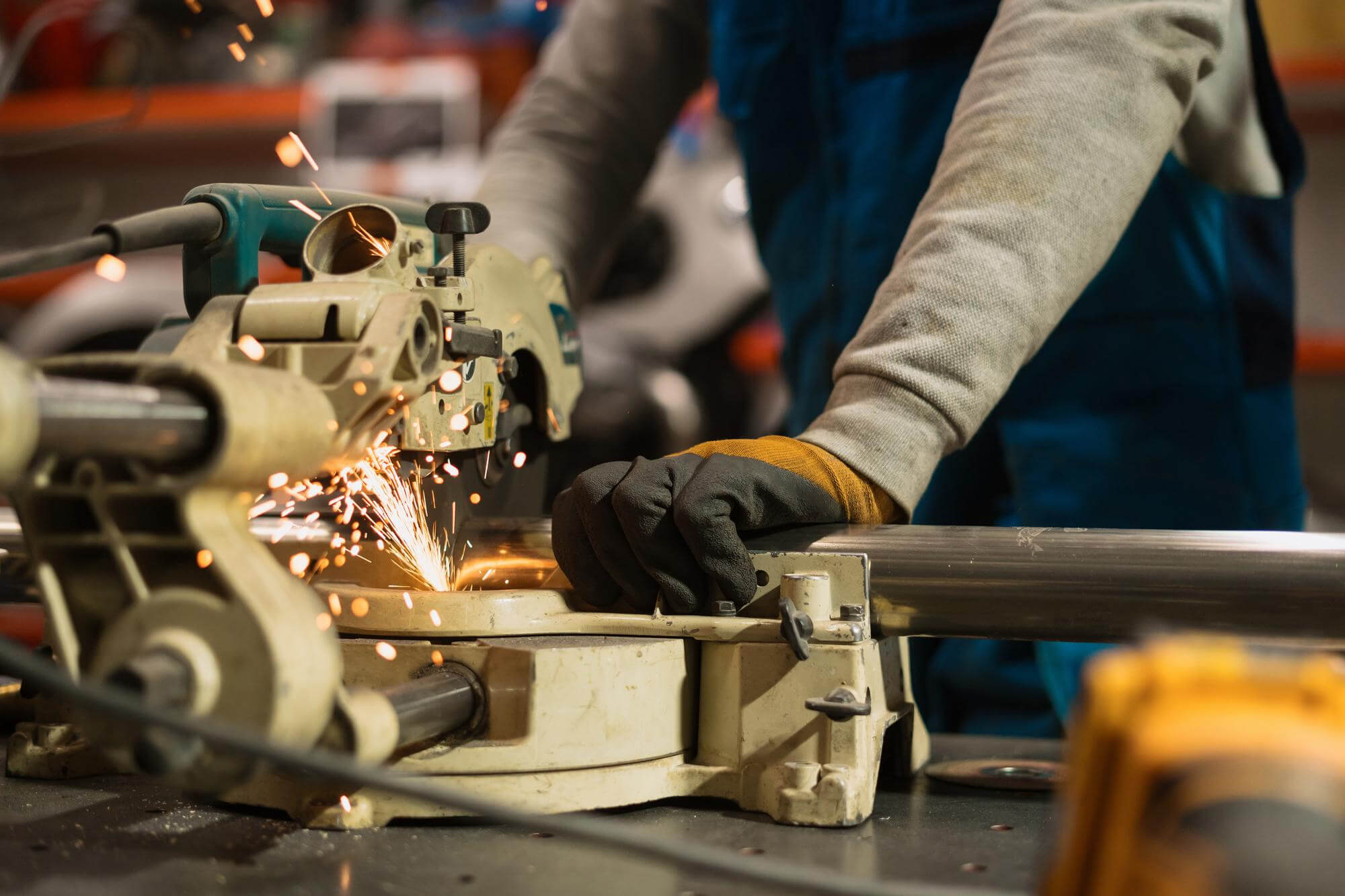 Worker using a metal chop saw to cut a steel pipe with sparks flying, wearing protective gloves and workwear.