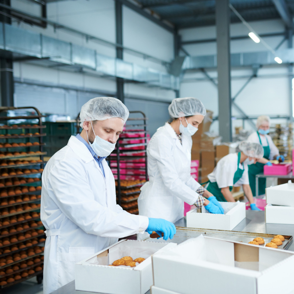 Workers in hairnets, face masks, and gloves packing baked cookies into white boxes in a bakery production line.