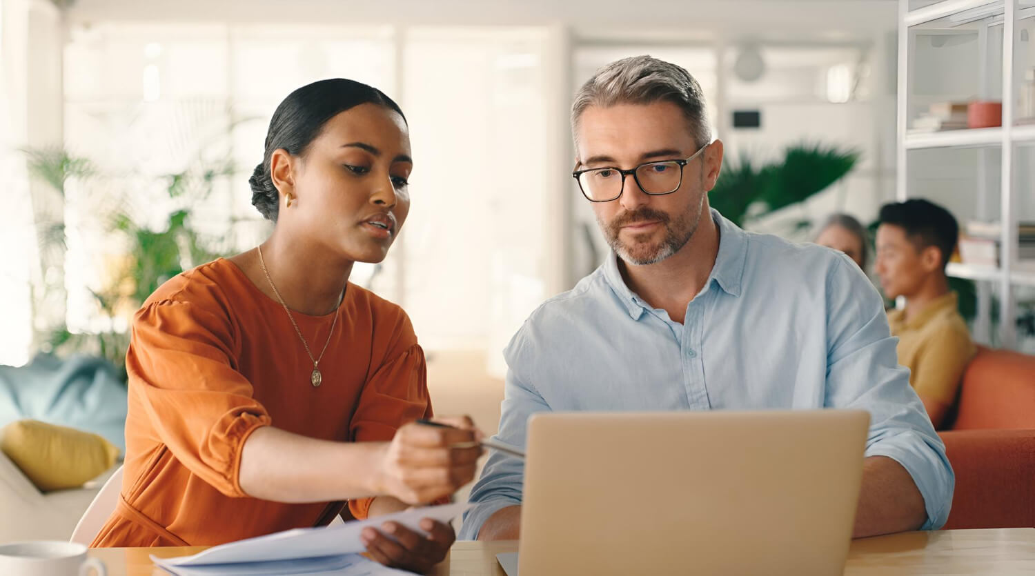 A woman in an orange blouse explaining documents to a man with glasses working on a laptop in a bright modern office.