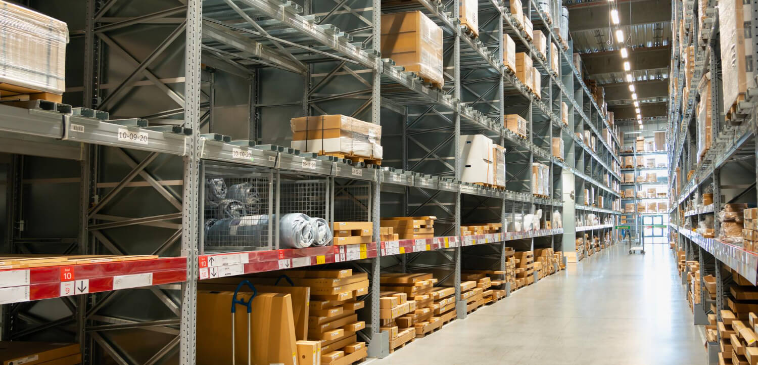 Wide aisle between tall metal shelves stocked with boxes and wrapped items in a large warehouse.