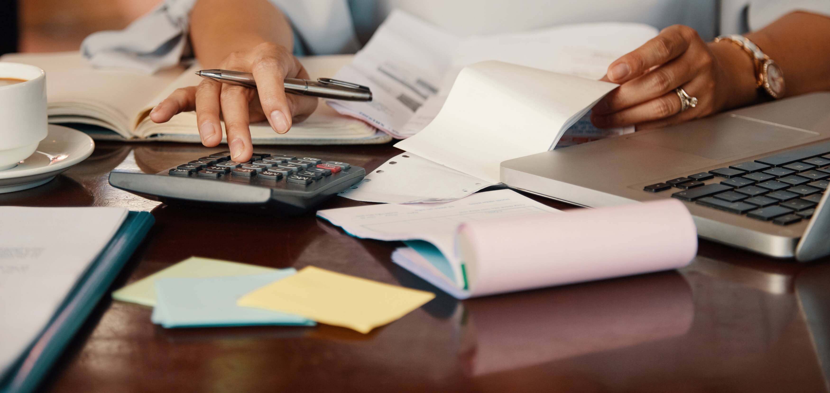 Person using calculator and reviewing documents next to a laptop and coffee cup on a desk.