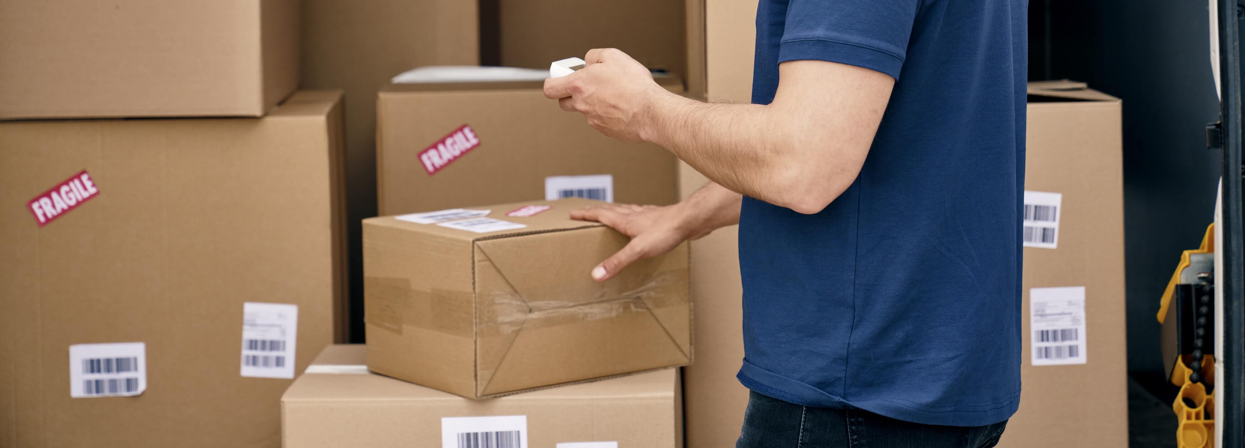 Person in blue shirt scanning barcodes on stacked cardboard boxes labeled fragile inside a delivery vehicle.