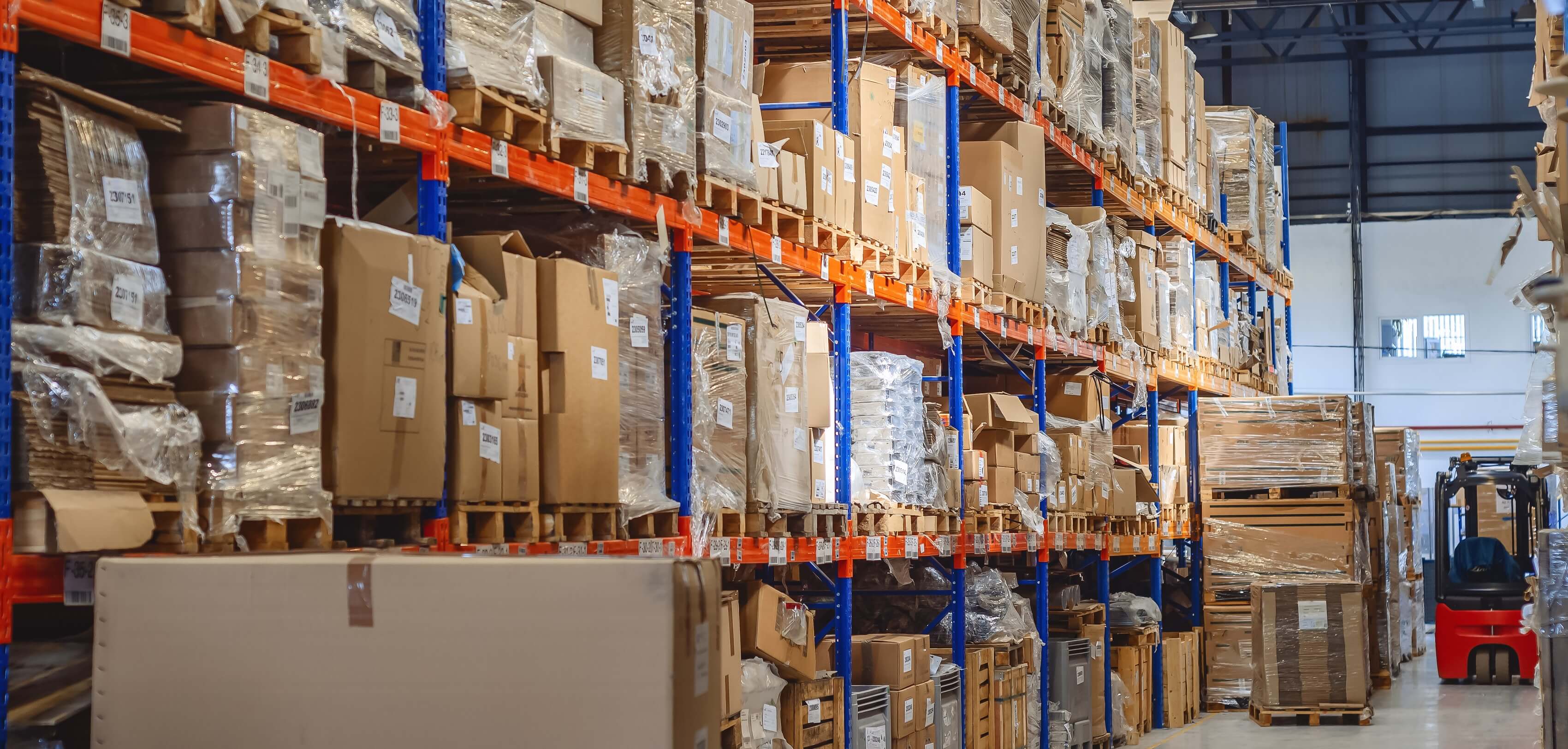 Warehouse storage shelves stacked with cardboard boxes and pallets, with a red forklift in the aisle.