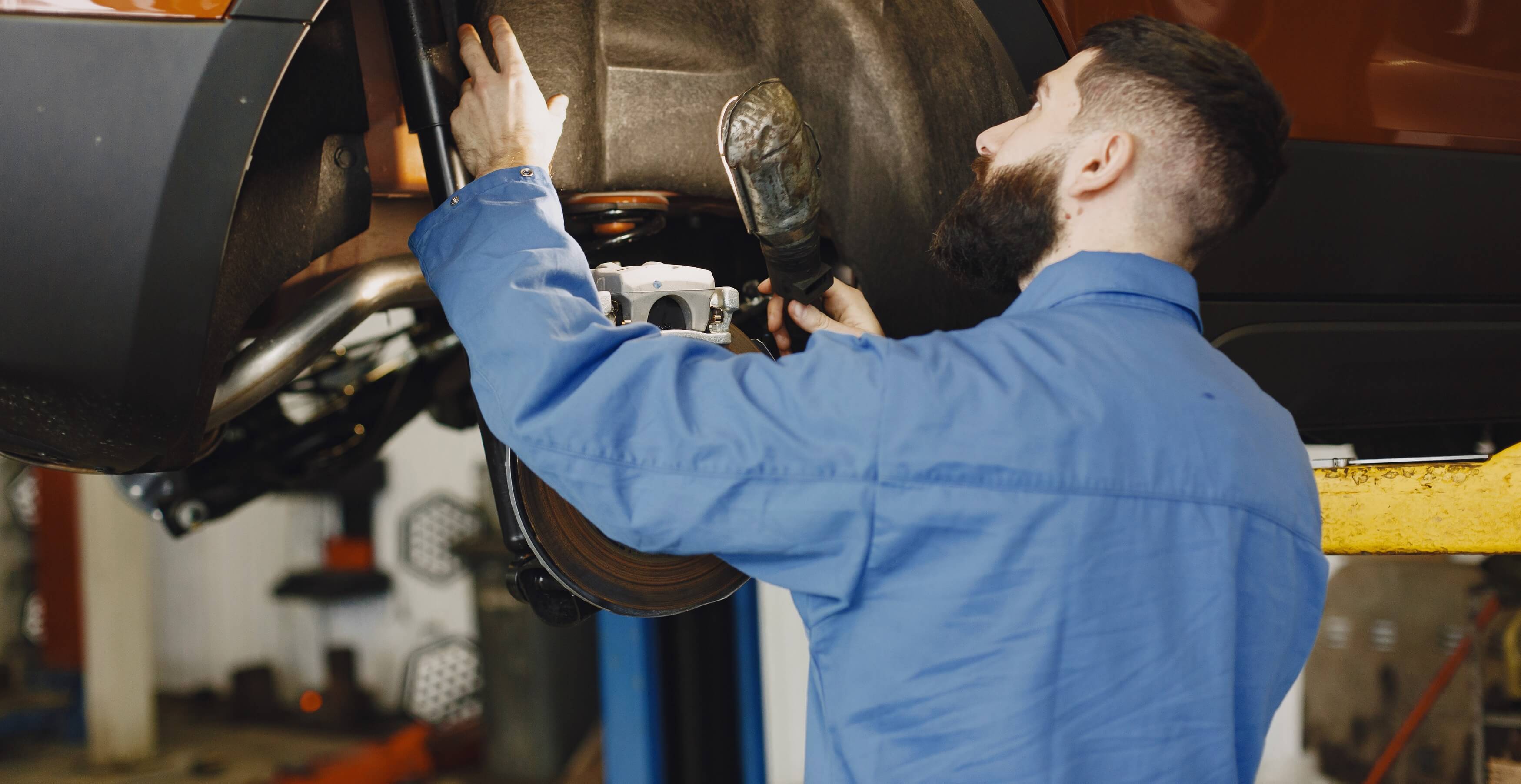 Bearded mechanic in blue coveralls inspecting the underside of an elevated orange vehicle in a garage.