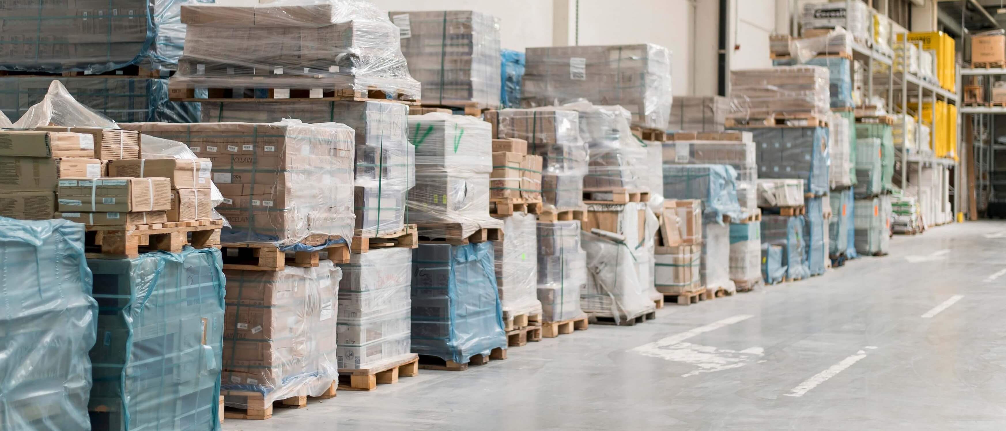 Warehouse interior with stacks of pallets wrapped in plastic containing various boxed goods along a wide aisle.