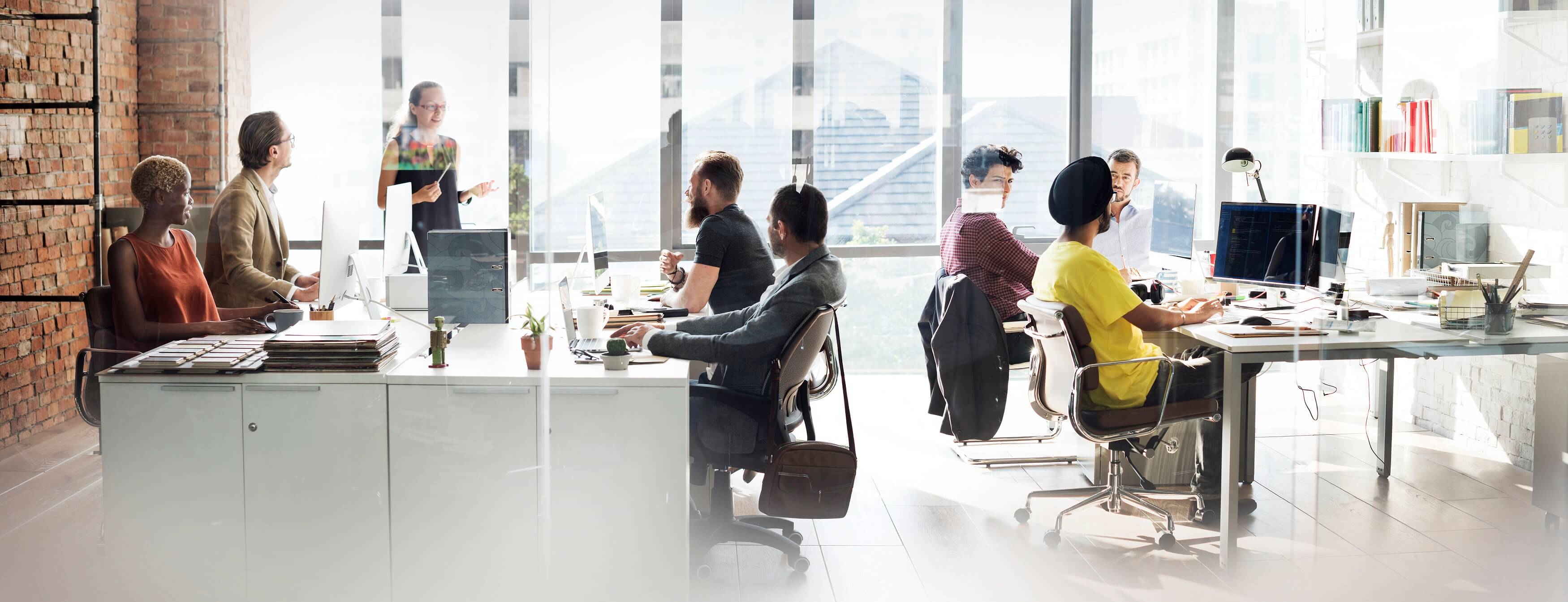 Diverse group of seven people collaborating and working on computers in a bright, modern office with large windows and exposed brick wall.