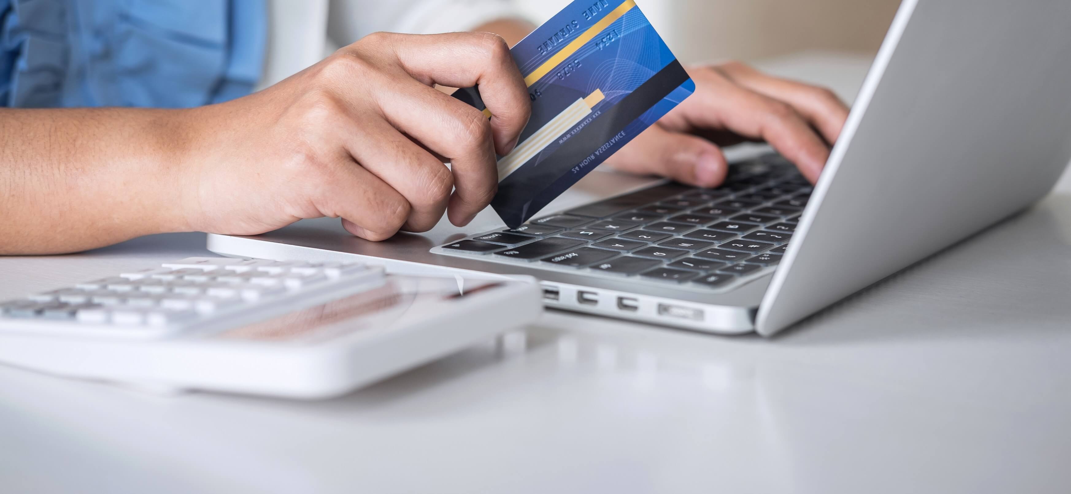 Person holding a credit card while typing on a laptop keyboard with a calculator on the desk.
